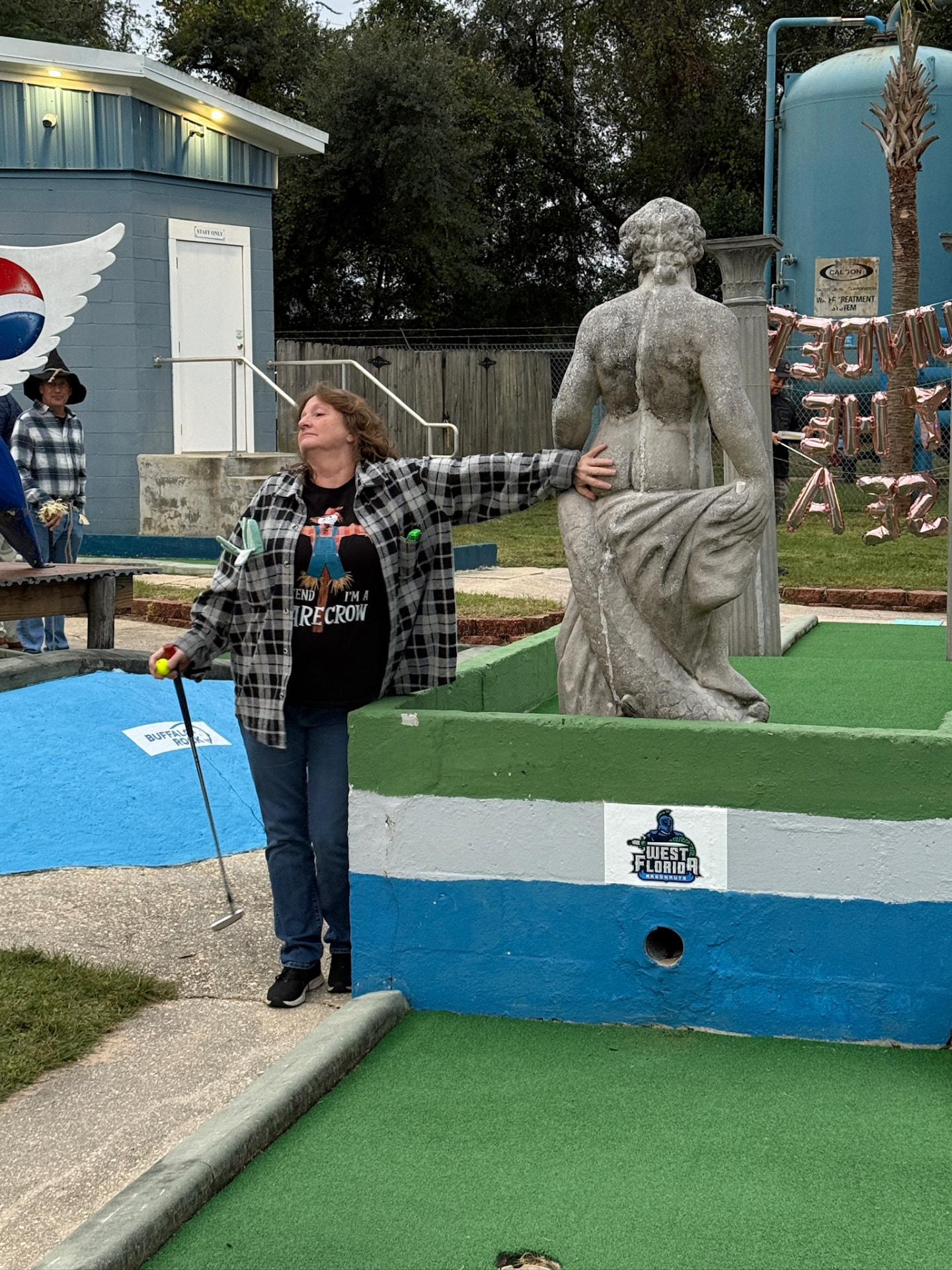 Player in a scarecrow shirt putting beside a Goofy Golf statue